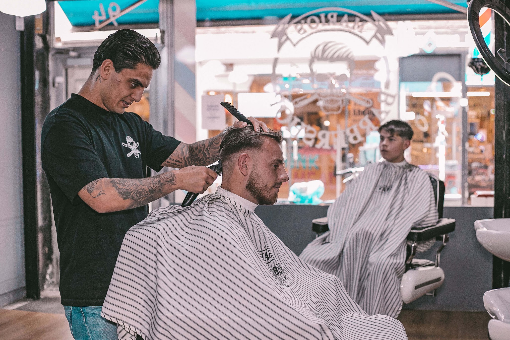 Een barbier in een zwart shirt met het logo knipt een klant, met de etalage en de straat op de achtergrond