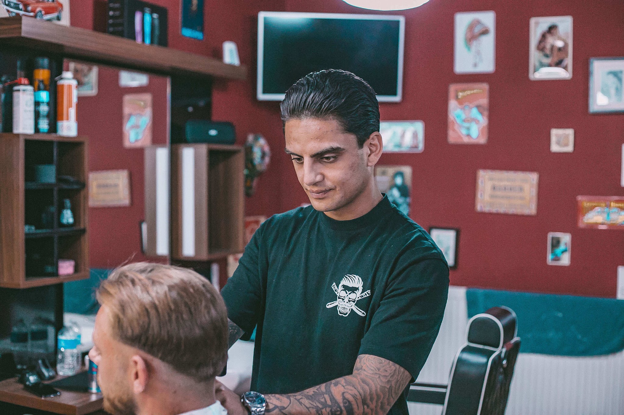 Een barbier in een groen shirt met het logo knipt een klant, met vintage bordjes aan de rode muur