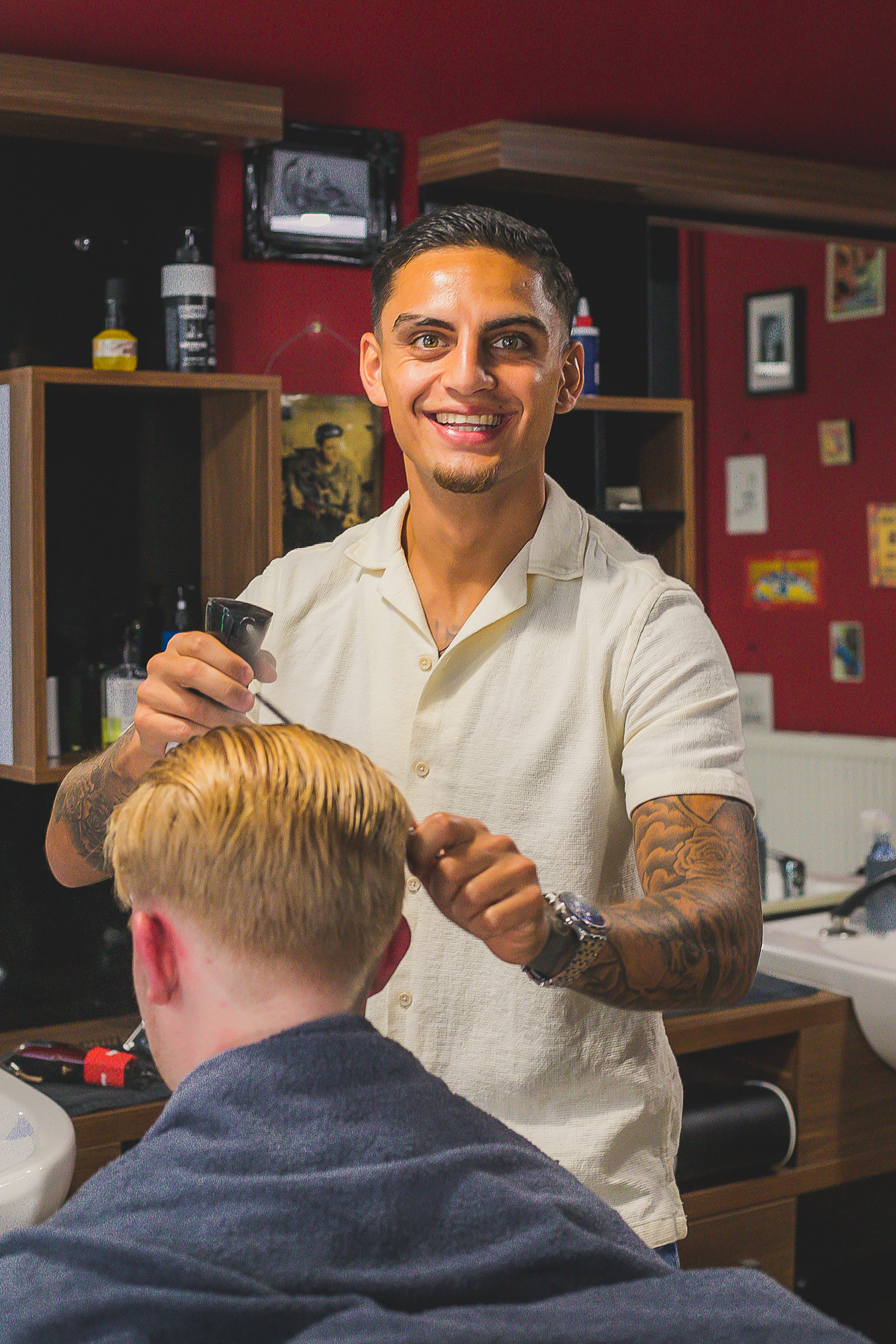 Een lachende barbier in een crèmekleurig overhemd knipt een klant voor de rode muur
